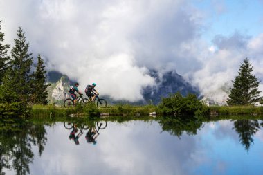 Brunni, İsviçre - Ağustos 06. 2021: İnsanlar su yüzeyinde refleksle Haerzlisee dağı boyunca dağ bisikletlerine biniyorlar. Titlis bölgesindeki Engelberg 'in yukarısındaki Haerzlisee bölgesi, farklı çeşitlilikler sunan popüler bir dinlenme yeridir.