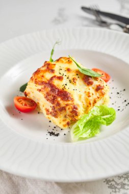 Domatesli lazanya ve yeşil yaprak. Beşamel soslu balık. Işık masasında beyaz restoran tabağı