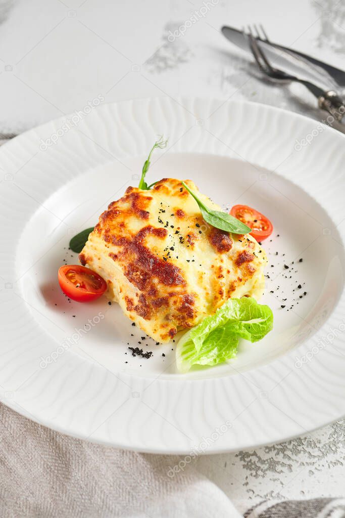 Lasagna with tomato and green leaf. Fish with bechamel sauce. White restaurant plate on light table