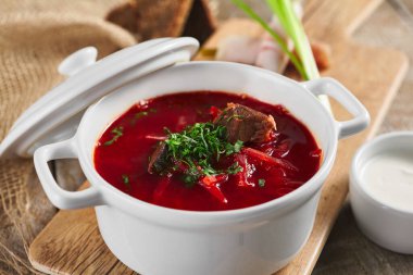 Bir kase kırmızı pancar kökü çorbası. Beyaz kâse içinde, ahşap masa üzerinde. Yeşil soğan, beyaz ekşi krema, ekmek ve sarımsak ile süslenmiş.