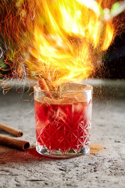 Fiero cocktail with whiskey and cinnamon stick with rosemary twig side view. Alcohol beverage close up with cinnamon powder. Night club drink with ice cubes in bar glassware on blurred background