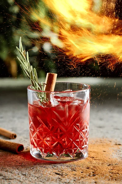 Fiero cocktail with whiskey and cinnamon stick with rosemary twig side view. Alcohol beverage close up with flame sparkles. Night club drink with ice cubes in bar glassware on blurred background