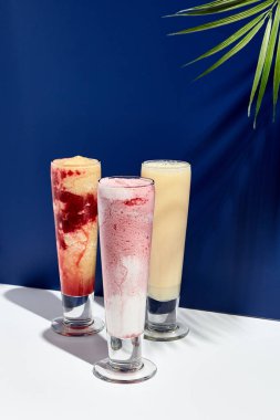 Ahududu ve muz, ananas, çilek ve hindistan cevizi sütü üzerine beyaz ve mavi arka plan. Gün ışığı ve sert gölgeli palmiye yaprağı. Yaz, sağlıklı, taze içecek konsepti