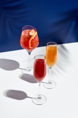 Aperol içeceği ve bellini kokteyli beyaz masada, mavi arka planda. Güneş ışığı ve palmiye yaprağı gölgesi modern natürmortu çeker. Yaz, parti, tropikal ve tazelik konsepti
