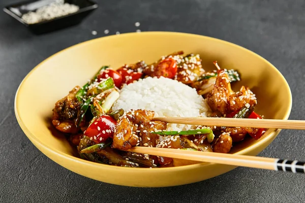 Sweet and Sour Chicken and Vegetables with Rice. Stir Fry Chicken with Vegetables and Sesame. Yellow plate with wooden chopstick on dark slate table. Chinese, asian, authentic food concept.