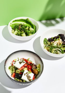 Yunan salatası, peynir ve pancar salatası ve chuka yosunlu salatalık. Yeşil duvarlı beyaz masada salata kasesi. Yeşil tropik yaprağın sert gölgesinde güneş ışığı