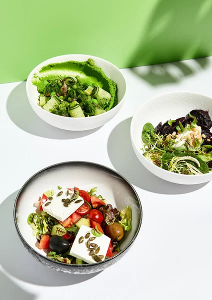 Yunan salatası, peynir ve pancar salatası ve chuka yosunlu salatalık. Yeşil duvarlı beyaz masada salata kasesi. Yeşil tropik yaprağın sert gölgesinde güneş ışığı