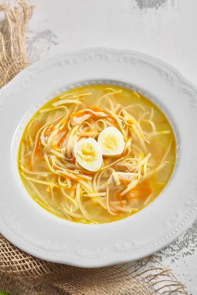 Chicken noodle soup with quail egg. Homestyle chicken noodle soup in white bowl on rustic white table with sack napkin
