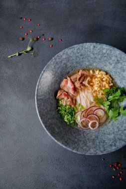 Vietnam mutfağı - Biftek Pho Erişte Çorbası veya Pho Bo Çorbası. Dokulu Koyu Gri Masa 'da Mavi Kasede Taze Yeşiller ile servis edildi. Baharatlı Pho Bo Çorbası. Üst Görünüm