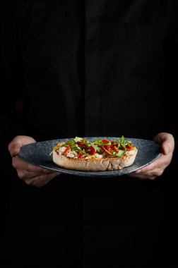 Şef İtalyan bruschetta tabağı tutuyor. Koyu mavi tabakta kurutulmuş domates ve Mozzarella peyniri. Arkaplanda Siyah Şef Üniforması