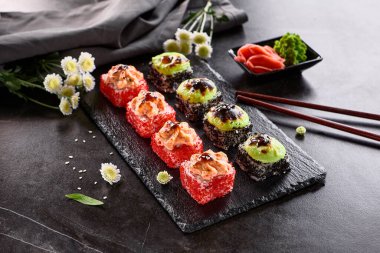 Uluslararası maki suşisi siyah tabakta yuvarlanıyor. Tahta çubuk, yeşil wasabi ve turşulu zencefil ile servis edilir. Japon seti, koyu taştan arka planda maki suşi rulosu..