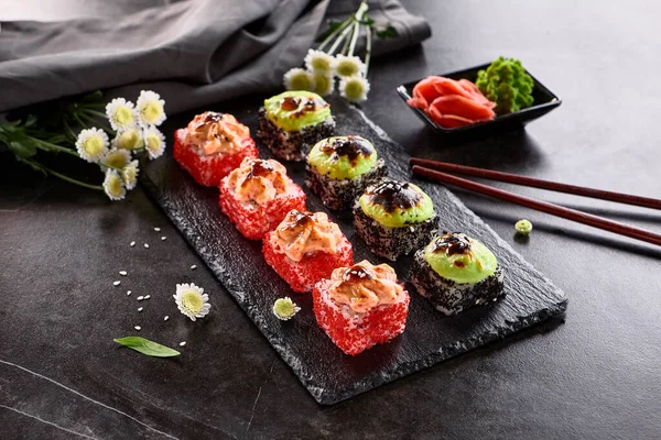 Uluslararası maki suşisi siyah tabakta yuvarlanıyor. Tahta çubuk, yeşil wasabi ve turşulu zencefil ile servis edilir. Japon seti, koyu taştan arka planda maki suşi rulosu..