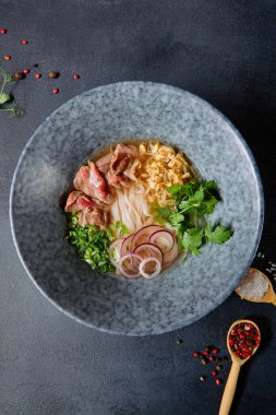 Vietnam mutfağı - Biftek Pho Erişte Çorbası veya Pho Bo Çorbası. Dokulu Koyu Gri Masa 'da Mavi Kasede Taze Yeşiller ile servis edildi. Baharatlı Pho Bo Çorbası. Üst Görünüm