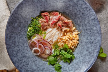 Vietnam mutfağı - Biftek Pho Erişte Çorbası veya Pho Bo Çorbası. Dokulu Koyu Gri Masa 'da Mavi Kasede Taze Yeşiller ile servis edildi. Baharatlı Pho Bo Çorbası. Üst Görünüm