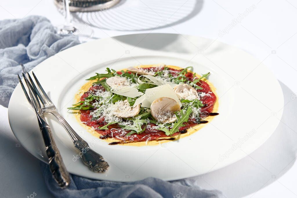 Beef carpaccio with mushrooms and parmesan cheese. Fancy dinning with beef carpaccio on white table with simple contemporary decor. Sunlight and harsh shadow still life. Beef carpaccio appetizer dish