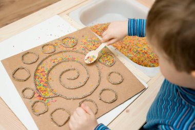 Çocuk renkli pirinçle oynuyor. Çocukların oyun yoluyla iyi motor becerileri geliştirmesi için uygulama. El-göz koordinasyonunu öğrenmek için oyuncak. Evde Montessori tipi anaokulu aktiviteleri.