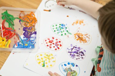 Evde Paskalya aktiviteleri. Karantina sırasında dalga geçin. Çocuk, yumurta şekillerini kulaklık ve guaj ile süslemiş. Evde DIY oyunları.