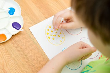 Evde Paskalya aktiviteleri. Karantina sırasında dalga geçin. Çocuk, yumurta şekillerini kulaklık ve guaj ile süslemiş. Evde DIY oyunları.