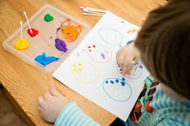 Evde Paskalya aktiviteleri. Karantina sırasında dalga geçin. Çocuk, yumurta şekillerini kulaklık ve guaj ile süslemiş. Evde DIY oyunları.