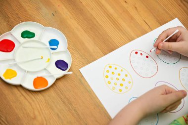 Evde Paskalya aktiviteleri. Karantina sırasında dalga geçin. Çocuk, yumurta şekillerini kulaklık ve guaj ile süslemiş. Evde DIY oyunları.