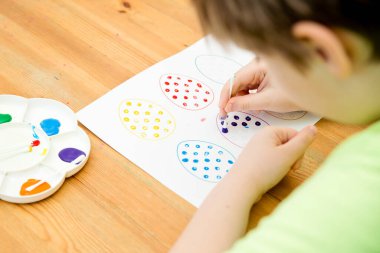 Evde Paskalya aktiviteleri. Karantina sırasında dalga geçin. Çocuk, yumurta şekillerini kulaklık ve guaj ile süslemiş. Evde DIY oyunları.