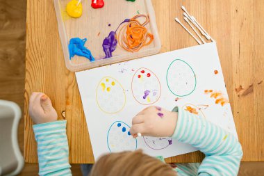 Evde Paskalya aktiviteleri. Karantina sırasında dalga geçin. Çocuk, yumurta şekillerini kulaklık ve guaj ile süslemiş. Evde DIY oyunları.