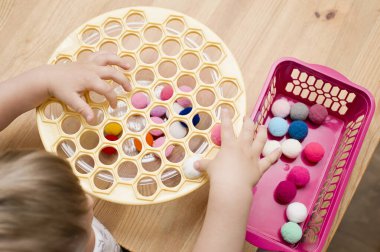 3 yaşında bir çocuk deliğe ponponları sokarak Rus köfte makinesiyle oynuyor. Plastik sepette renkli iplik topları