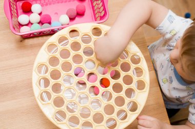 3 yaşında bir çocuk deliğe ponponları sokarak Rus köfte makinesiyle oynuyor. Plastik sepette renkli iplik topları