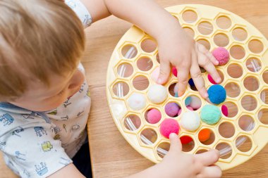 3 yaşında bir çocuk deliğe ponponları sokarak Rus köfte makinesiyle oynuyor. Plastik sepette renkli iplik topları
