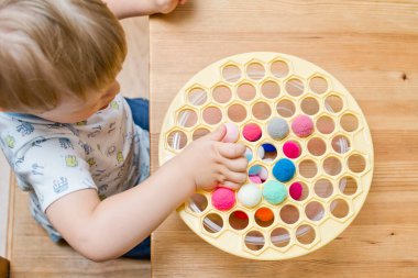 3 yaşında bir çocuk deliğe ponponları sokarak Rus köfte makinesiyle oynuyor. Plastik sepette renkli iplik topları