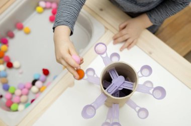 Evindeymiş gibi davran. Koordinasyon, denge ve motor becerileri geliştirmek için DIY oyuncağı. Çocuk, tatlı kaşıklarına ponpon yerleştiriyor..