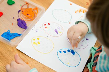 Evde Paskalya aktiviteleri. Karantina sırasında dalga geçin. Çocuk, yumurta şekillerini kulaklık ve guaj ile süslemiş. Evde DIY oyunları.