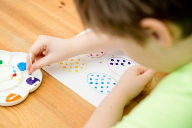 Evde Paskalya aktiviteleri. Karantina sırasında dalga geçin. Çocuk, yumurta şekillerini kulaklık ve guaj ile süslemiş. Evde DIY oyunları.