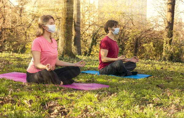 Young couple in gymsuit with Mask and Closed Eyes Sitting on Yoga Mat in Relaxing Meditation Pose at Park with Sun Reflections.