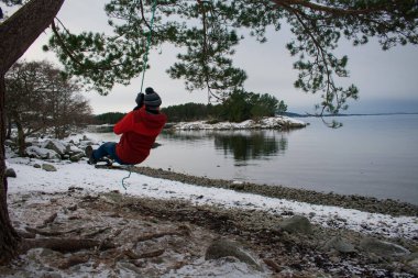 Sıcak giysili genç bir adam, donmuş gölün kenarında sallanan bir salıncak. Kış tatili konsept resmi.