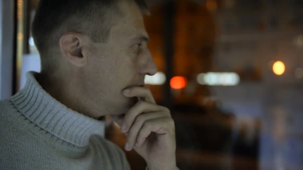 Депрессивный человек рядом с окном на фоне вечерней улицы, автомобилей и огней — стоковое видео