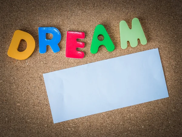 Palavra de madeira colorida DREAM com pedaço de papel branco sobre placa de cortiça — Fotografia de Stock