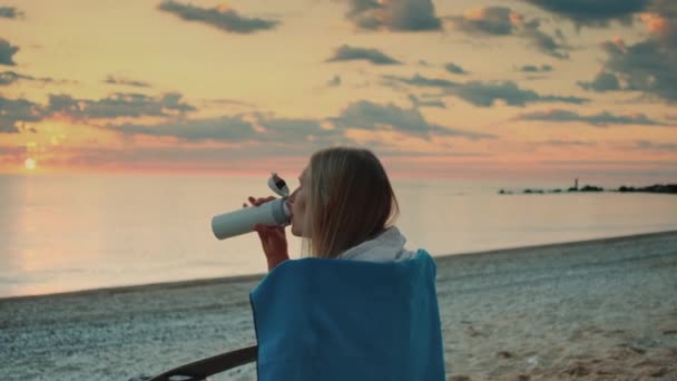 Jeune femme buvant dans une tasse thermique et assise sur la plage avant le lever du soleil — Video