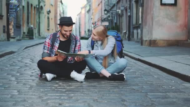 Young tourists couple using map, sitting on pavement and admiring historical surroundings — Stock Video