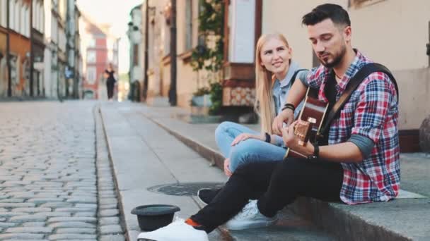 Jonge stel straatzangers zitten op de stoep, spelen gitaar en zingen — Stockvideo
