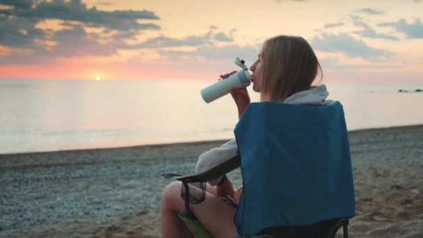 Young woman drinking from thermos and sitting on camping chairs on the beach — Stock Video