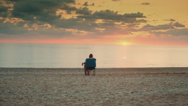 Vista da senhora admirando o pôr do sol no mar sentado na cadeira turística dobrável — Vídeo de Stock