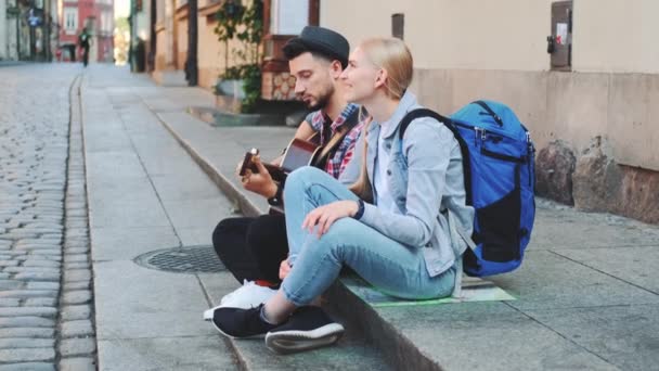 Пара туристів сидить на тротуарі, грає на гітарі і відпочиває — стокове відео