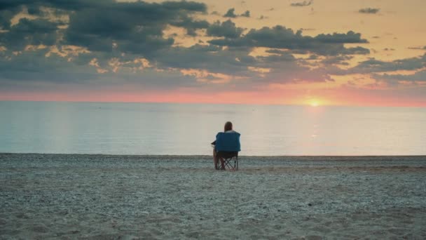 Deniz kenarında gün batımını seyreden bir kadın. Katlanan turist koltuğunda oturuyor. — Stok video