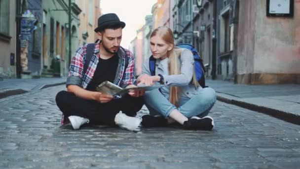 Pareja de turistas jóvenes usando el mapa, sentados en el pavimento y admirando los alrededores históricos — Vídeo de stock