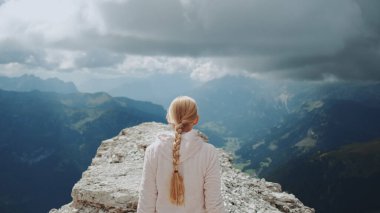 Back view of blonde woman walking on the top of the mountain under the clouds clipart