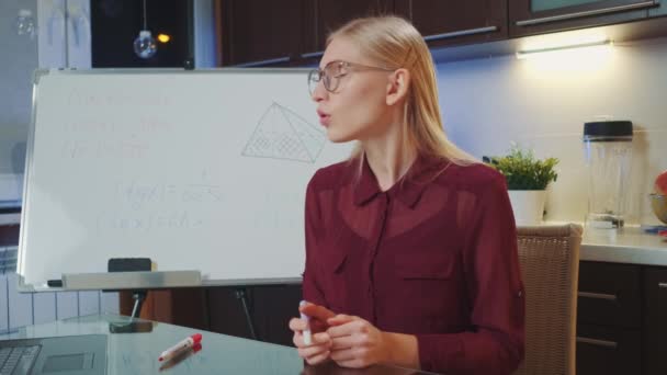 Mujer rubia de negocios con gafas de vista hablando con la cámara y escribiendo en pizarra — Vídeo de stock