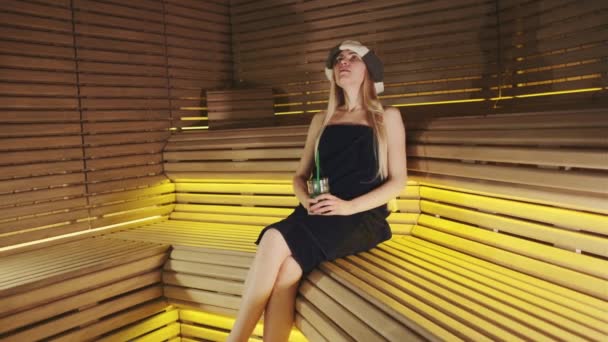 Mujer rubia bonita en sombrero de sauna y toalla de baño sentado y sintiéndose bien en sauna — Vídeo de stock