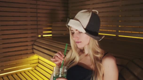 Szőke nő szauna kalapban, szalmaszálból iszik üdítőt. — Stock videók
