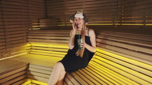 Fiatal nő szaunában beszél okostelefonnal és kezében üdítőital — Stock videók